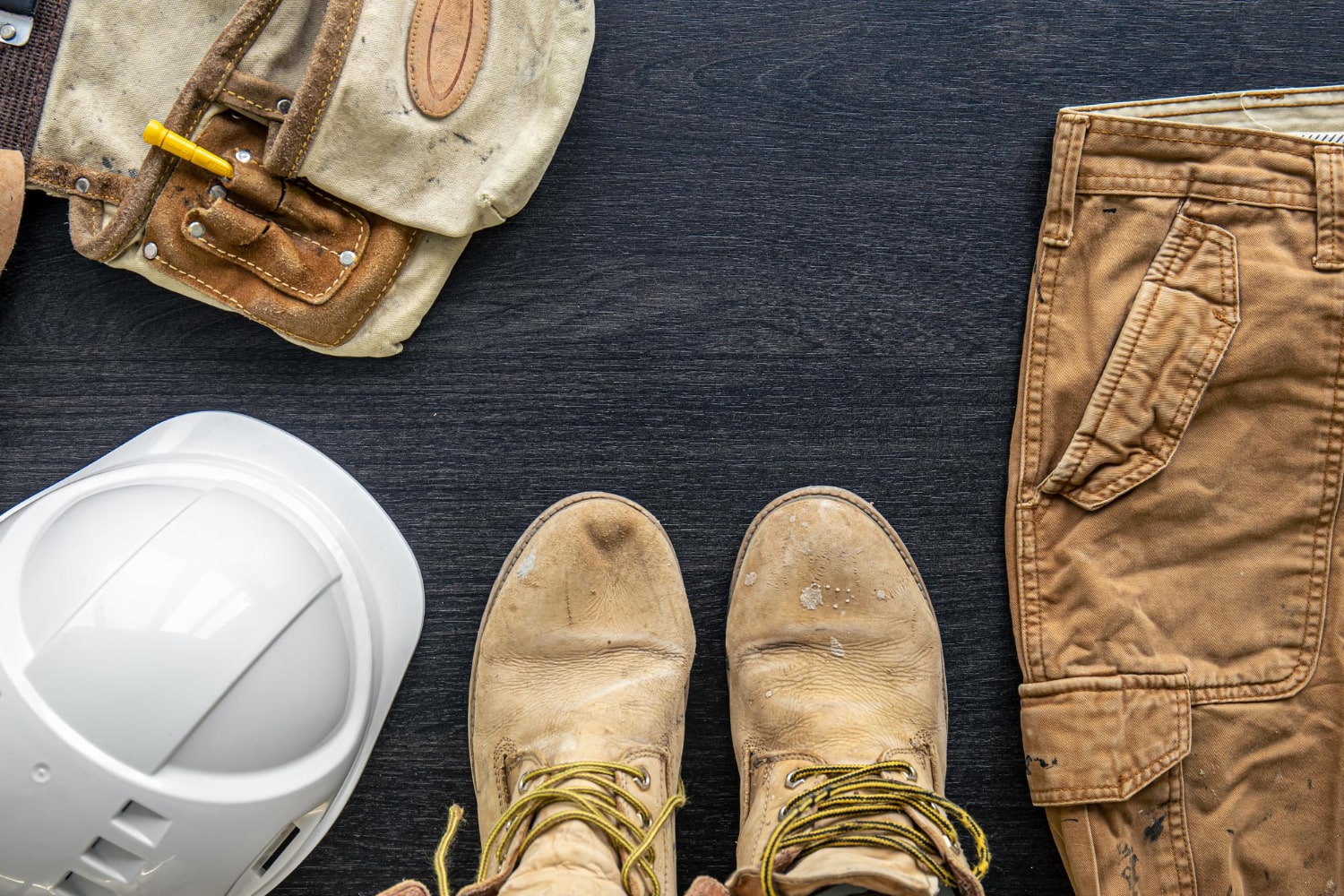 Viršutinis vaizdas, kuriame matomi darbiniai batai, apsauginis šalmas, kelnės ir įrankių diržas, išdėstyti ant tamsaus paviršiaus